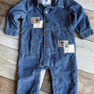 以下が含まれることがあります： 青色のコーデュロイベビーロンパース。襟付きでボタン留め、2つの花柄の装飾パッチと襟に小さなラベルが付いています。長袖で、脚にはスナップが付いています。