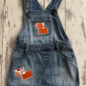 Puede incluir: Vestido peto vaquero azul claro con un bolsillo en el pecho y dos bolsillos delanteros. El vestido presenta parches florales y naranjas en el peto y la falda. Las correas son ajustables y se aseguran con botones.