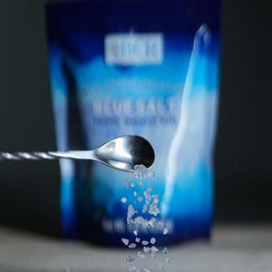 May include: A close-up shot of blue salt crystals being poured from a spoon. A blue package with the text "BLUE SALT" and the "BCR" logo is in the background. The salt is on a wooden surface.