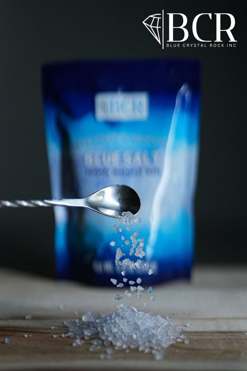 May include: A close-up shot of blue salt crystals being poured from a spoon. A blue package with the text "BLUE SALT" and the "BCR" logo is in the background. The salt is on a wooden surface.