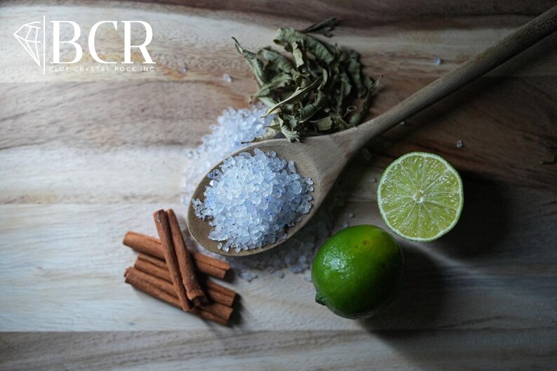 May include: A wooden spoon filled with blue-tinted salt sits on a wooden surface, next to a lime, cinnamon sticks, and dried herbs. The image includes the text "BCR BLUE CRYSTAL ROCK INC."