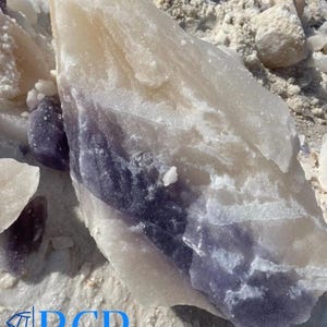 May include: A large, raw crystal formation with a layered appearance. The stone features bands of white, cream, and deep purple hues. The image also shows smaller fragments and the logo "BCR BLUE CRYSTAL ROCK INC."
