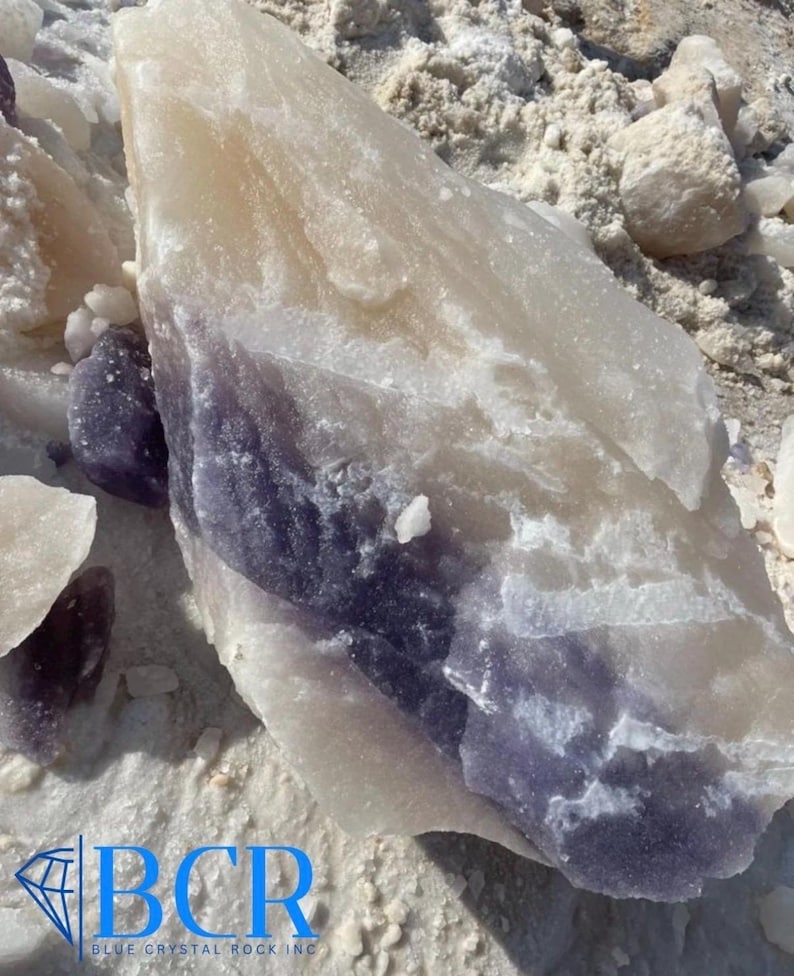 May include: A large, raw crystal formation with a layered appearance. The stone features bands of white, cream, and deep purple hues. The image also shows smaller fragments and the logo "BCR BLUE CRYSTAL ROCK INC."