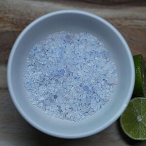 May include: A white ceramic bowl filled with coarse, light blue and white bath salts. Two lime halves are on the right side of the bowl, on a wooden surface. The bath salts have a textured appearance.