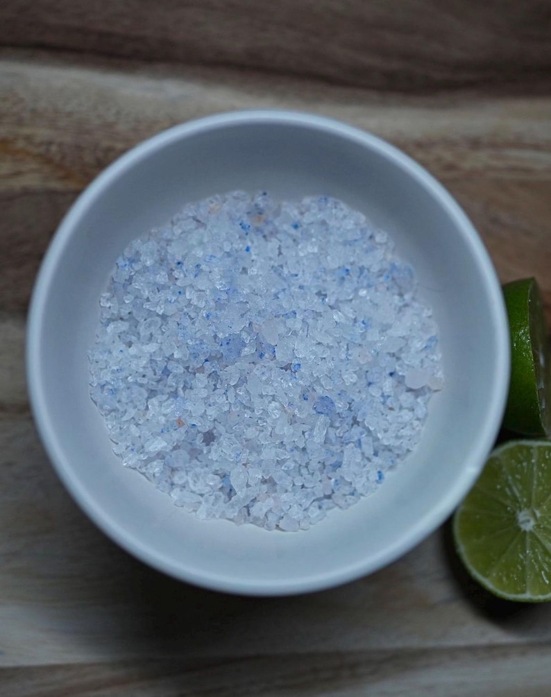 May include: A white ceramic bowl filled with coarse, light blue and white bath salts. Two lime halves are on the right side of the bowl, on a wooden surface. The bath salts have a textured appearance.