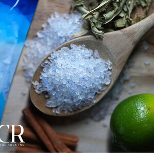 May include: A wooden spoon filled with blue-tinged salt crystals, with a bag of salt in the background. Limes, cinnamon sticks, and dried herbs are also visible. The image is a close-up shot of the ingredients.
