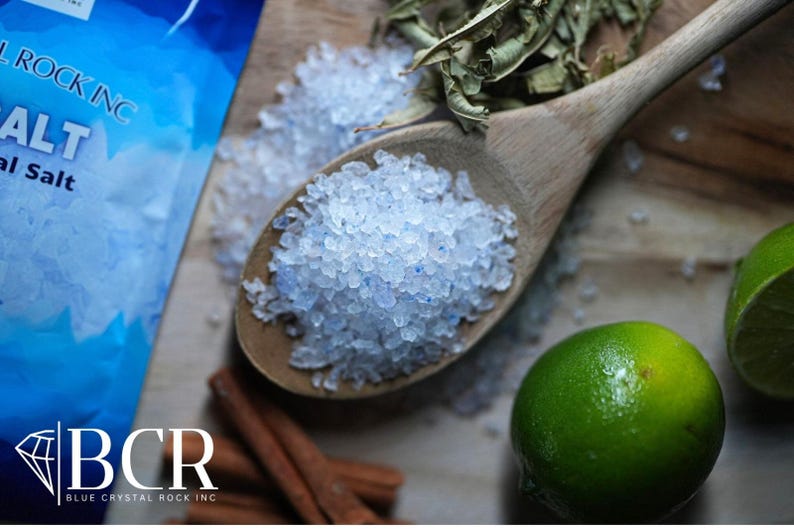 May include: A wooden spoon filled with blue-tinged salt crystals, with a bag of salt in the background. Limes, cinnamon sticks, and dried herbs are also visible. The image is a close-up shot of the ingredients.