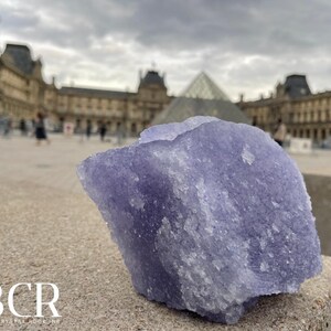 May include: A large, rough-hewn, lavender-colored crystal is the focal point of this image. The crystal sits on a stone surface, with the Louvre Museum and a cloudy sky in the background. The crystal has a textured surface and a natural, organic shape.
