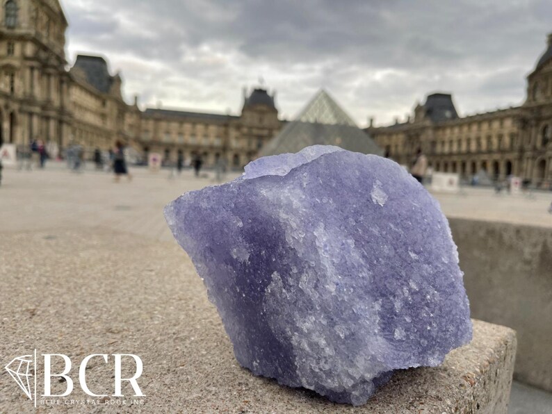 May include: A large, rough-hewn, lavender-colored crystal is the focal point of this image. The crystal sits on a stone surface, with the Louvre Museum and a cloudy sky in the background. The crystal has a textured surface and a natural, organic shape.