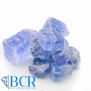 May include: A pile of rough, blue crystal rocks against a white background. The crystals vary in size and shape, with a translucent quality. The logo "BCR BLUE CRYSTAL ROCK INC" is in the lower left corner.