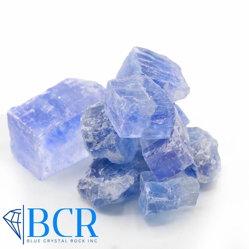 May include: A pile of rough, blue crystal rocks against a white background. The crystals vary in size and shape, with a translucent quality. The logo "BCR BLUE CRYSTAL ROCK INC" is in the lower left corner.