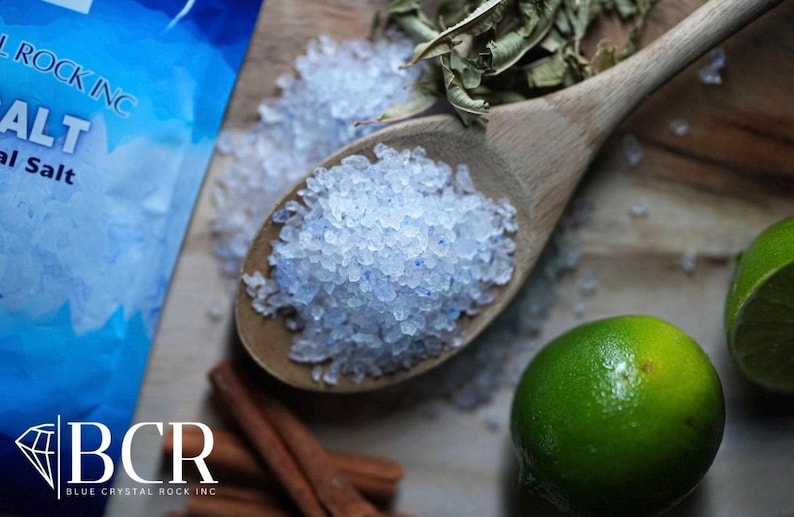 May include: A wooden spoon filled with blue-tinted salt crystals, alongside a bag of salt, fresh limes, dried herbs, and cinnamon sticks. The image showcases ingredients for culinary or spa use, with a focus on natural elements.