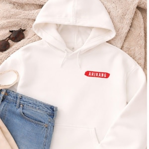 May include: A white hooded sweatshirt with a kangaroo pocket and drawstring. The word "ARIRANG" is embroidered in red on the chest. Sunglasses and blue jeans are visible in the image.