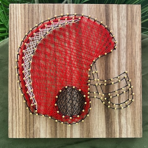 Puede incluir: Obra de arte de hilo hecha a mano que representa un casco de fútbol americano. El casco está hecho con hilo rojo, negro, blanco y dorado sobre un fondo de madera. El diseño presenta una representación detallada de la forma del casco.