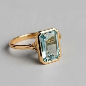 May include: A gold ring with a rectangular, pale blue gemstone. The ring has a delicate band and a bezel setting. The gemstone is a light, translucent blue, and the ring is set against a neutral background. The ring's design is simple and elegant.