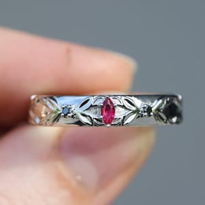 May include: A silver ring with a central marquise-cut ruby and floral engravings. The ring features a delicate design with small black gemstones and is held between fingers. A second ring is visible in the background.