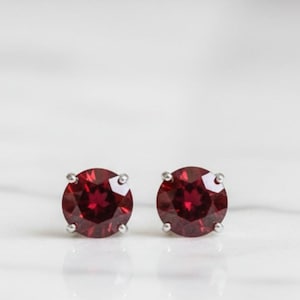 Peut inclure: Une paire de boucles d'oreilles à clous avec des pierres précieuses rondes, rouge foncé, serties dans du métal argenté. Les boucles d'oreilles sont présentées sur une surface en marbre blanc, mettant en valeur leur couleur vibrante et leur design simple.