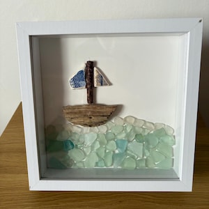 May include: A framed artwork depicting a sailboat made from driftwood and ceramic, sailing on a sea of sea glass. The sea glass is in shades of blue and green. The artwork is presented in a white shadow box frame.