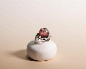 May include: A silver ring with a large, round, reddish-brown gemstone. The ring is set on a white, rounded pedestal against a neutral background. The gemstone has a speckled appearance.