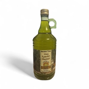 May include: A clear glass bottle of olive oil with a handle and a gold cap. The label displays "Ancient Tree" in English and Arabic. The olive oil has a light green colour. The bottle is approximately 30 cm tall.