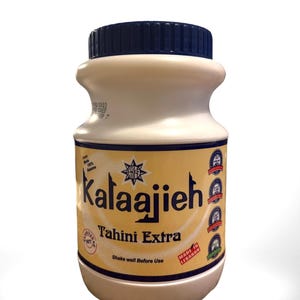 May include: A cylindrical jar of Kalaajieh Tahini Extra. The jar is off-white with a dark blue lid. The label is tan with the brand name in large, stylized letters. Text on the label includes "Tahini Extra" and "Shake well before use."