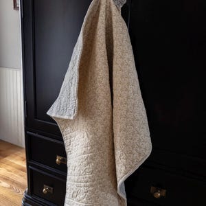 May include: A beige and gray quilted blanket with a floral pattern hangs on a black cabinet. The blanket has a light gray border and is folded over, showcasing the detailed stitching and design. The cabinet has brass hardware.