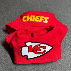 Puede incluir: Un conjunto rojo para mu&ntilde;eca, compuesto por una camisa y una visera. La camisa muestra un logotipo de flecha blanco y rojo con una "K" estilizada, y la visera tiene la palabra "CHIEFS" en letras amarillas.
