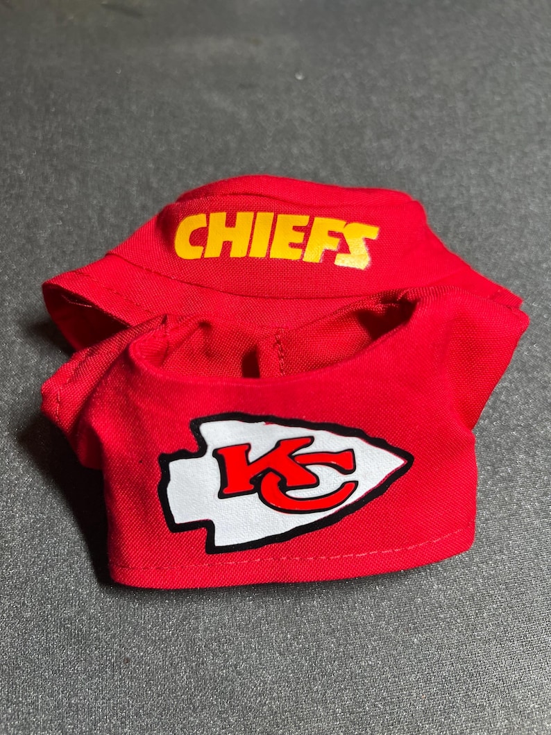 Puede incluir: Un conjunto rojo para mu&ntilde;eca, compuesto por una camisa y una visera. La camisa muestra un logotipo de flecha blanco y rojo con una "K" estilizada, y la visera tiene la palabra "CHIEFS" en letras amarillas.