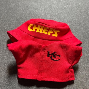 Puede incluir: Conjunto rojo de gorro de pescador y camisa. El gorro tiene la palabra "CHIEFS" en letras amarillas. La camisa tiene un logotipo "KC" negro. El conjunto es de tela suave.