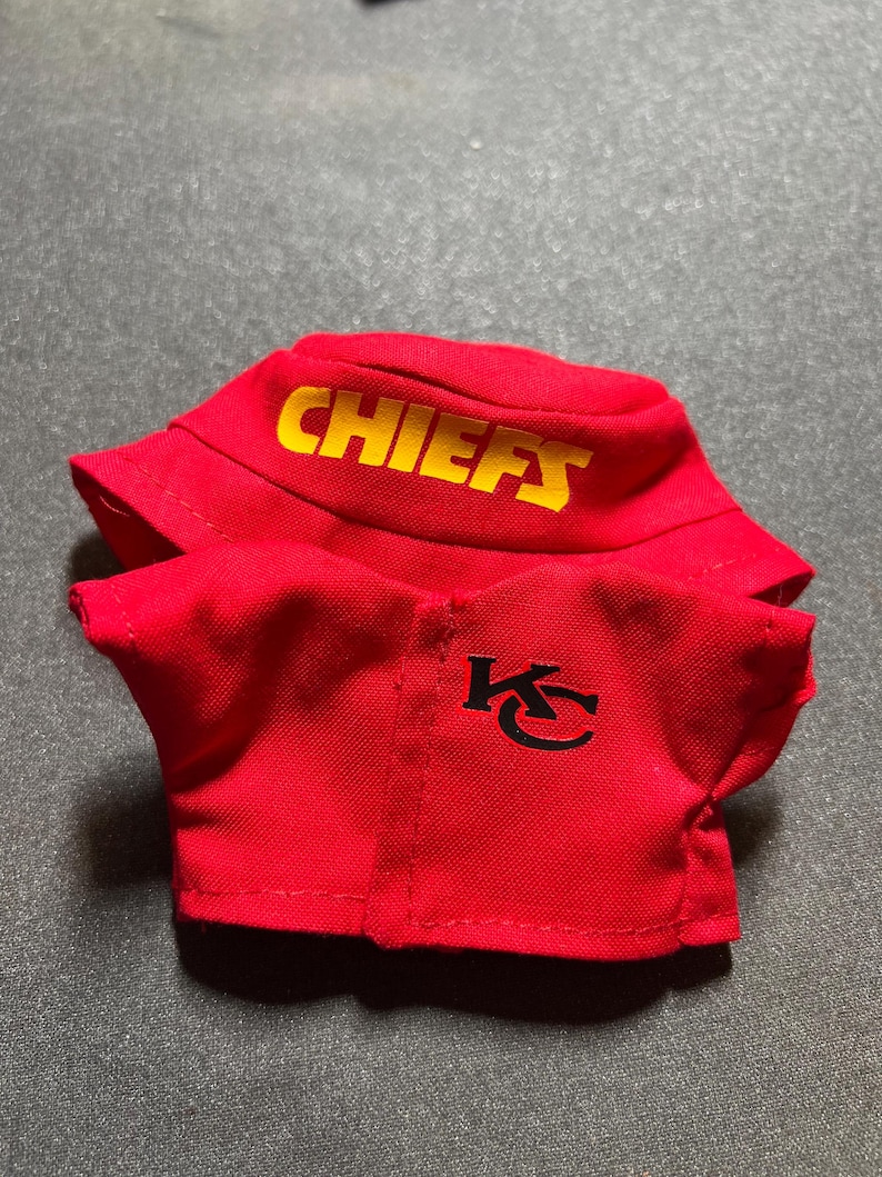 Puede incluir: Conjunto rojo de gorro de pescador y camisa. El gorro tiene la palabra "CHIEFS" en letras amarillas. La camisa tiene un logotipo "KC" negro. El conjunto es de tela suave.