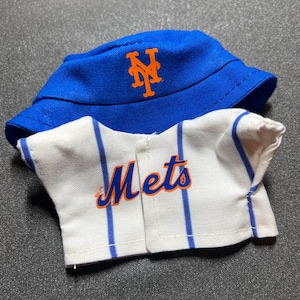May include: A blue bucket hat with an orange "NY" logo and a white baseball jersey with blue stripes and the word "Mets" in orange script. The hat and jersey are likely for a child.