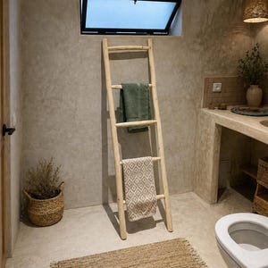 Puede incluir: Escena de baño con una escalera de madera apoyada en una pared de hormigón, sosteniendo toallas. Una toalla verde y otra estampada cuelgan de la escalera. Una alfombra tejida y una cesta con una planta están en el suelo. Una ventana abierta.
