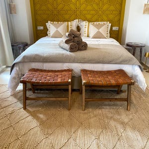 Könnte beinhalten: Ein Schlafzimmer mit einem Bett mit weißer Bettwäsche, gemusterten Kissen und einer braunen Decke. Zwei Bänke mit geflochtenen Ledersitzen stehen vor dem Bett auf einem rautenförmigen Teppich. Das Kopfteil ist senfgelb mit einem geometrischen Muster.