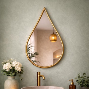 Puede incluir: Un espejo en forma de lágrima con un marco dorado cuelga de una pared verde claro. El espejo refleja una habitación con una lámpara colgante. Un jarrón con flores blancas, un grifo dorado y un dispensador de jabón están en primer plano.