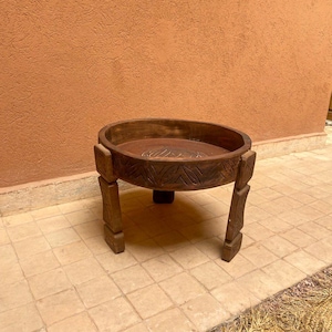 Peut inclure: Table en bois brun foncé, fabriquée à la main, avec un plateau rond et des détails sculptés. La table a quatre pieds robustes. Elle est posée sur un sol carrelé. Le design de la table suggère un style rustique ou bohème.
