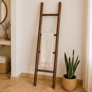 Puede incluir: Una escalera de madera marrón oscuro para toallas con una toalla crema colgada. Una planta de serpiente en maceta en una cesta de mimbre está al lado. La escena es en un baño con una paleta de colores neutros.