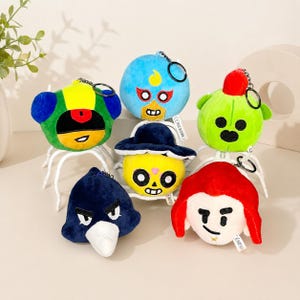 May include: A collection of six plush character keychains featuring various designs including a blue masked figure, a green cactus-like shape, a dark blue bird-like head, a yellow skull-faced character with a hat, and a red-haired figure.