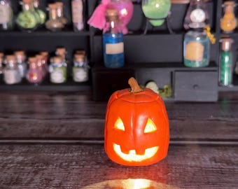 Può includere: Una zucca arancione intagliata con un volto sorridente e un interno luminoso. La zucca è il punto focale, con una superficie di legno in primo piano e una collezione di bottiglie di pozioni sullo sfondo. Decorazioni di Halloween.