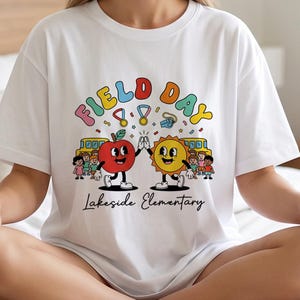 Pode incluir: Uma t-shirt branca com um gráfico colorido com o texto "FIELD DAY" e "Lakeside Elementary". O design inclui uma maçã e um sol de desenho animado a bater palmas, rodeados por crianças, autocarros escolares amarelos, medalhas e um apito.
