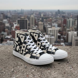 May include: Pair of high-top sneakers with a black and white abstract pattern. The sneakers have white laces, a white toe cap, and a white sole. The background features a cityscape.