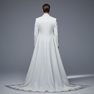 May include: A white formal coat with a long train. The coat has a tailored fit with a high collar and long sleeves. The train is decorated with a detailed, dark gray embroidered border. The person is facing away from the camera.