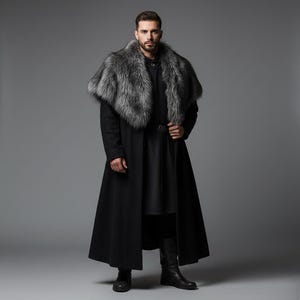 Op de afbeelding: Een lange zwarte jas met een grijze bontkraag. De jas heeft een riem in de taille en wordt gedragen met zwarte laarzen. De persoon staat voor een grijze achtergrond. De jas is in middeleeuwse stijl.