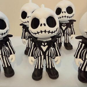 May include: Several figurines of Jack Skellington from The Nightmare Before Christmas. Each figure has a white head with black facial features, a black and white striped suit, and black shoes. The figures are standing on a white surface.
