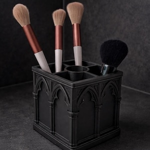 May include: A black makeup brush holder with a gothic arch design, holding four makeup brushes. Three brushes have light brown bristles, copper-colored handles, and white bases. The fourth brush has black bristles.