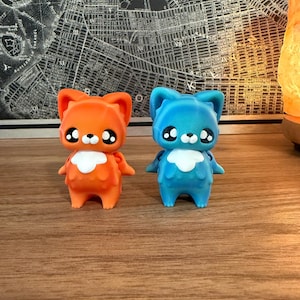 May include: Two small, cartoonish cat figurines. One is orange, the other is blue. Both have large, expressive eyes, white chest markings, and small wings. They are standing on a wooden surface.