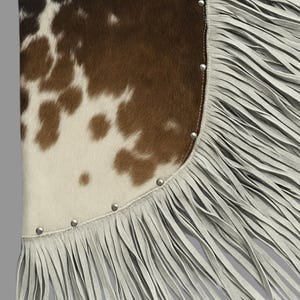 May include: A cowhide leather item with a brown and white spotted pattern. The edge is trimmed with light gray fringe and silver-colored studs.
