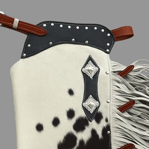 May include: Close-up of a western-style chaps. The chaps feature a cowhide pattern with black spots on a white background. The top is black leather with silver studs. Brown leather straps and white fringe complete the design.