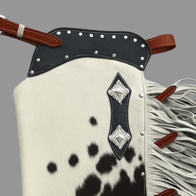 May include: Close-up of a western-style chaps. The chaps feature a cowhide pattern with black spots on a white background. The top is black leather with silver studs. Brown leather straps and white fringe complete the design.