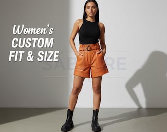 Op de afbeelding: Een vrouw draagt een zwarte top en een oranje leren short met een riem. De short heeft zakken en wordt gecombineerd met zwarte laarzen. De tekst "Women's Custom Fit & Size" en "Premium Quality Cowhide" is inbegrepen.