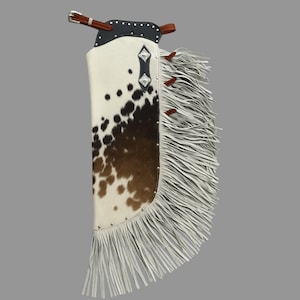 May include: Cowhide chaps with a white and brown spotted pattern. The chaps feature a black leather top with silver studs and a brown leather belt. White fringe adorns the side and bottom edges, adding a decorative element to the western-style garment.